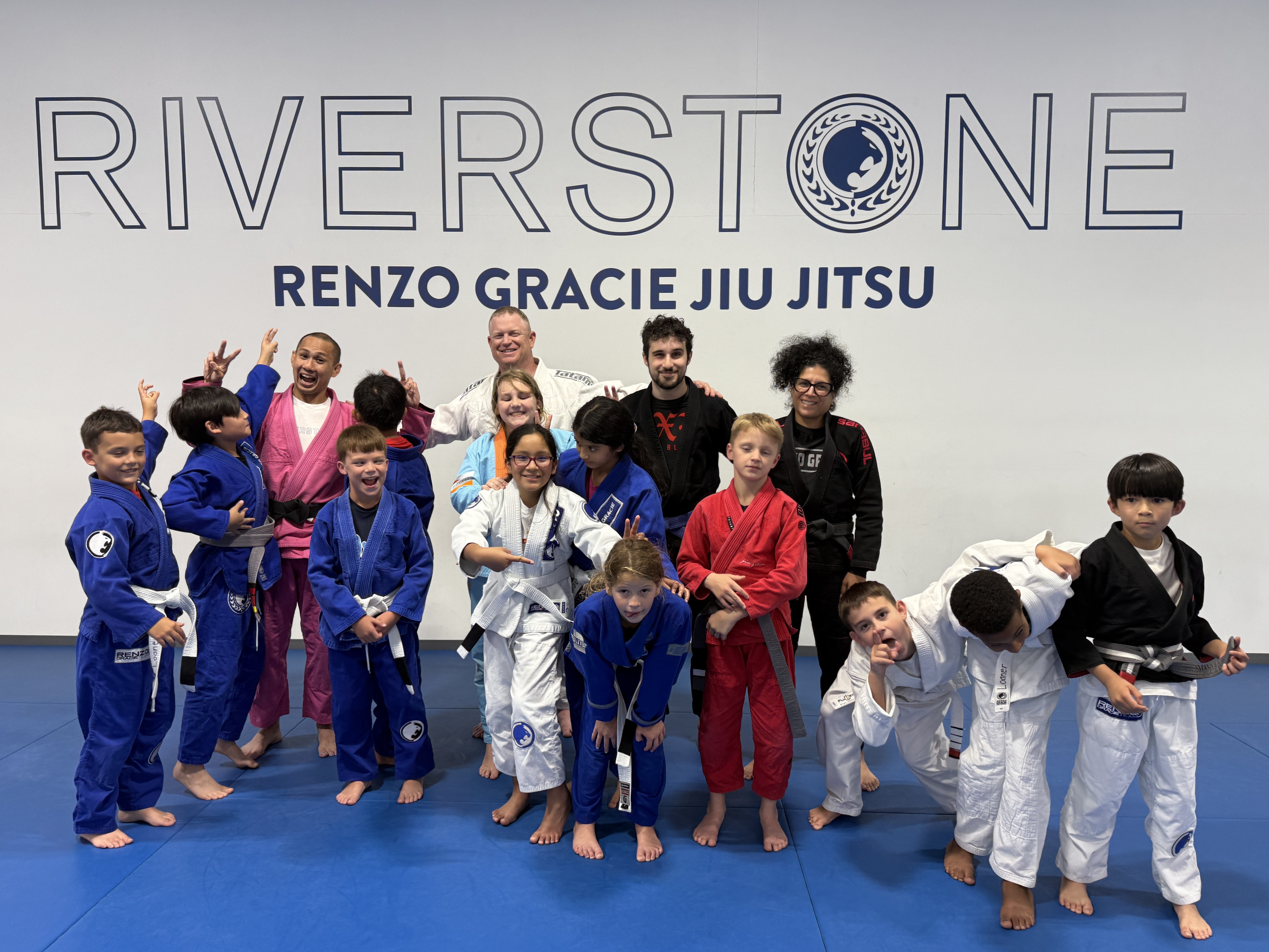 Group of children and instructors in Brazilian Jiu-Jitsu gis posing in front of the Riverstone Renzo Gracie Jiu Jitsu academy wall after class