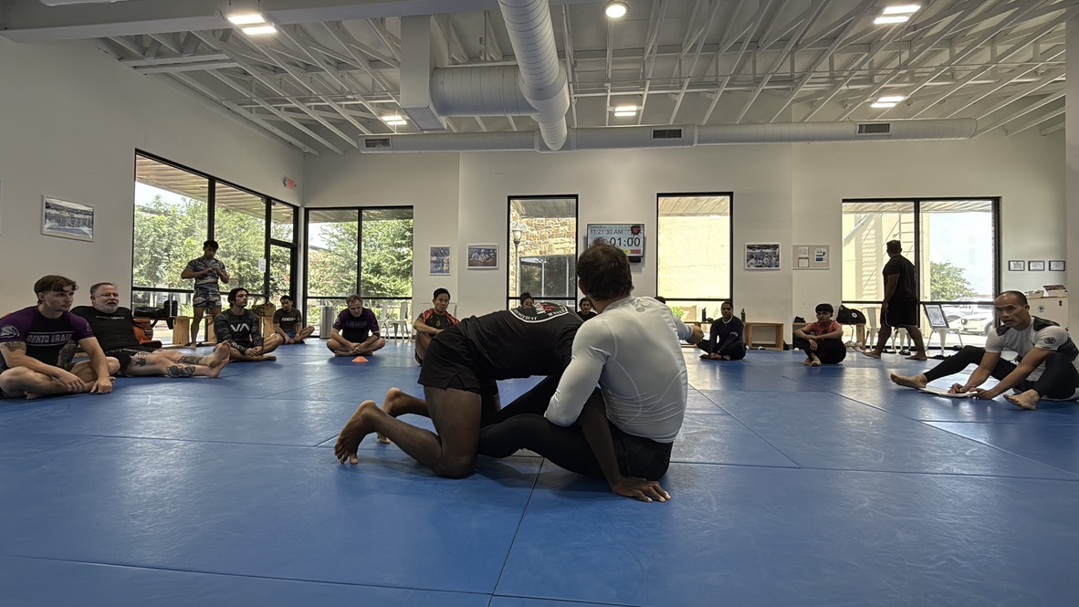 Brazilian Jiu-Jitsu class at Renzo Gracie Riverstone, students sitting on blue mats watching two practitioners drilling techniques, with natural light coming through large windows.