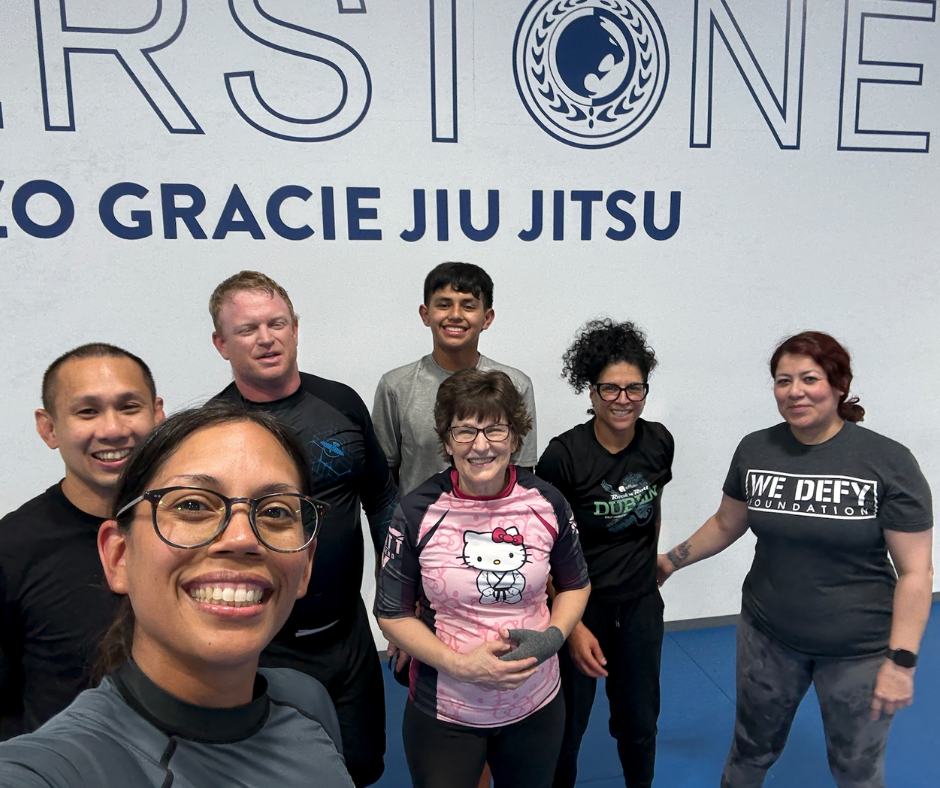 Smiling Brazilian Jiu-Jitsu students of all ages posing together after class at Renzo Gracie Riverstone, wearing training gear in front of academy wall.