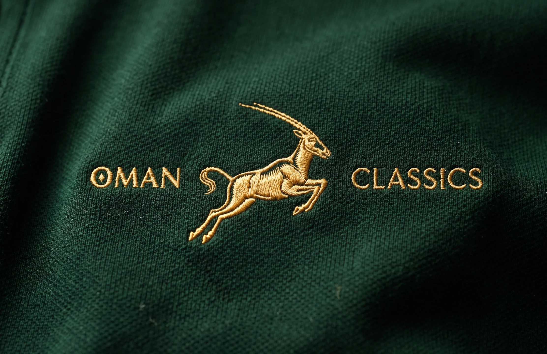 The Oman Classics logo embroidered in gold thread on a dark green polo shirt.