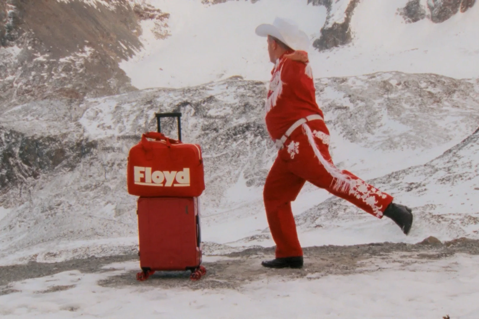 A cowboy in a red suit dancing past a red Floyd suitcase in the snowy mountains.