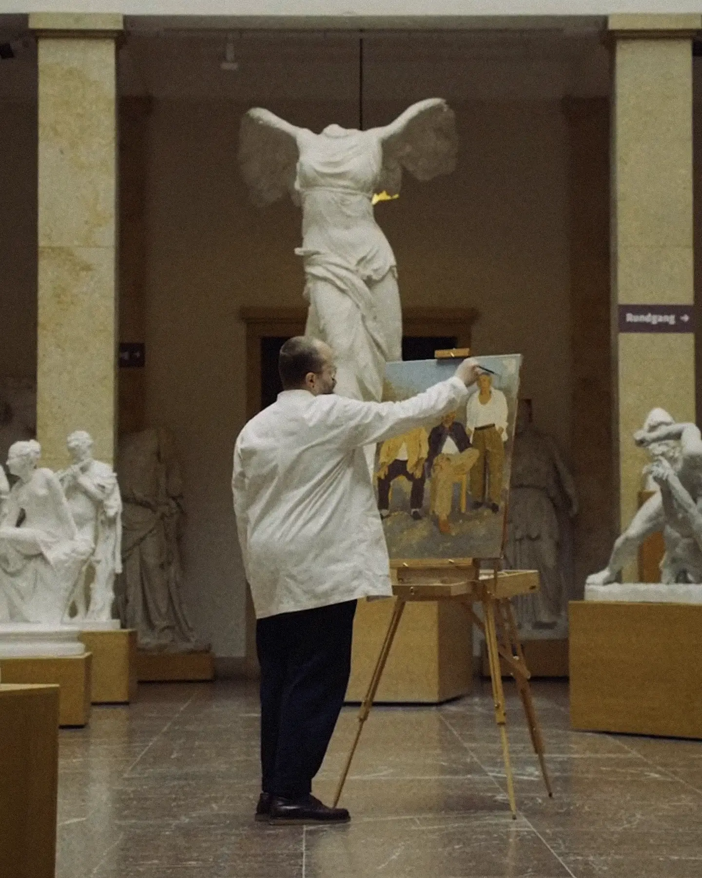 Constantin Spinner painting at an easel beneath a plaster cast of the Winged Victory of Samothrace, Museum of Casts, Munich — A Kind Of Guise campaign teaser by Studio ONOM.
