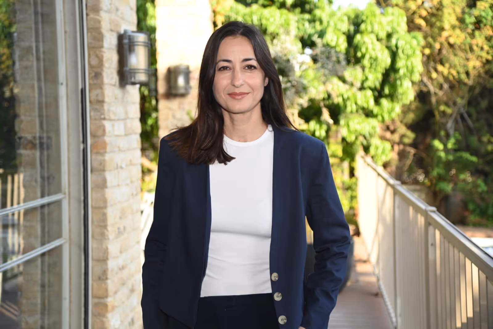 עינב חיים סיג, לבושה ז’קט כחול כהה וחולצה לבנה, מחייכת ועומדת מול רקע ירוק של צמחייה