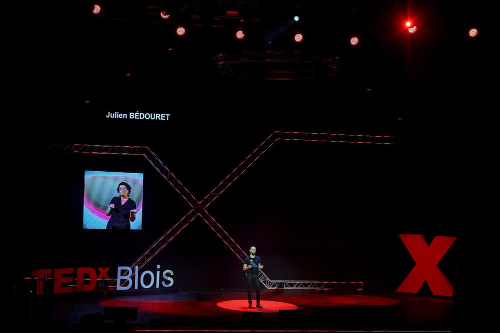 Scène du TEDx Blois 2021 sur laquelle parle Julien Bédouret du sujet de l'IA et du développement durable. Il y a aussi un écran pour retranscrire en langue des signes.