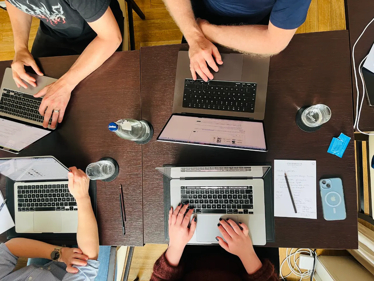 Vogelperspektive auf Arbeitsplatz mit vier Personen, die an Laptops arbeiten, mit Wassergläsern, Notizen und Smartphone auf dunklem Holztisch