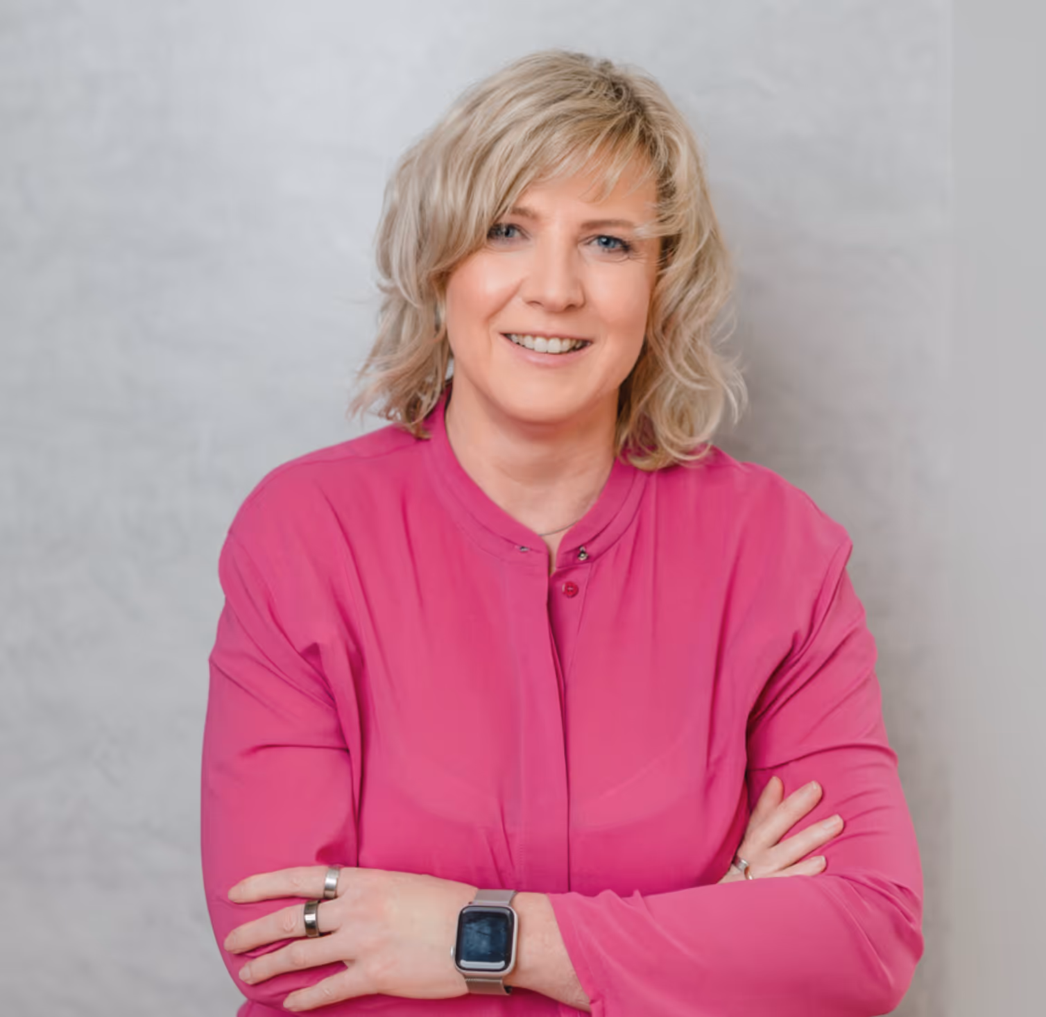 Ines Scholz mit blonden Haaren und pinker Bluse lächelt und verschränkt die Arme vor einem hellen Hintergrund.