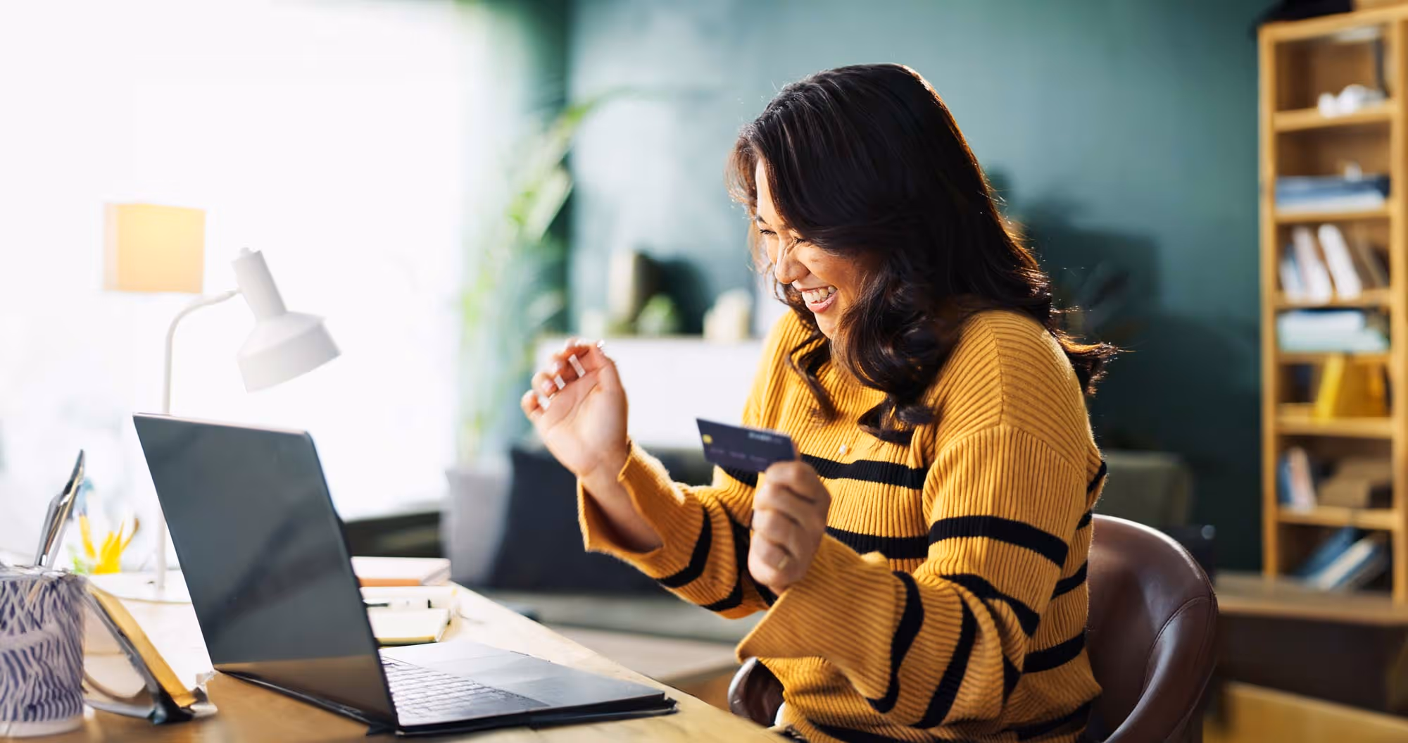 A woman smiles excitedly while holding her credit card and looking at her laptop.