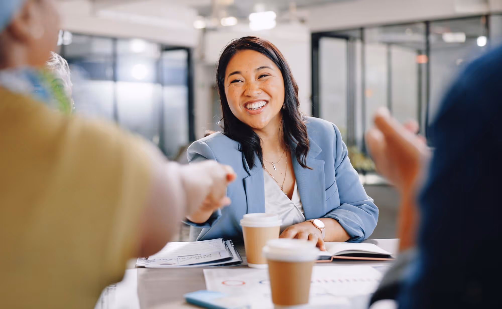 A young woman smiles and shakes hands with someone across the table in an office setting.