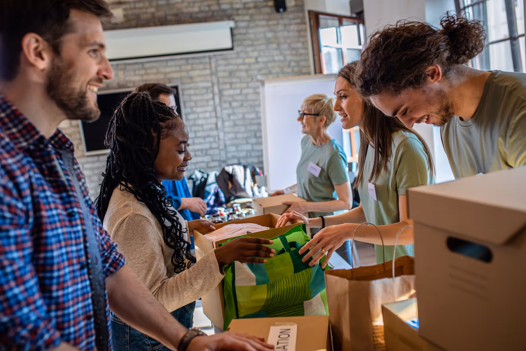 A diverse group of volunteers smiling and packing donation boxes and bags in a well-lit room with brick walls.