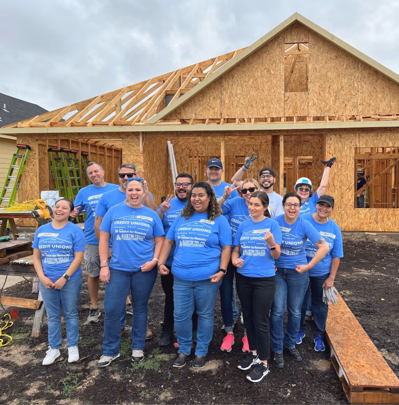 Group of smiling volunteers wearing blue shirts standing in front of a wooden house under construction.