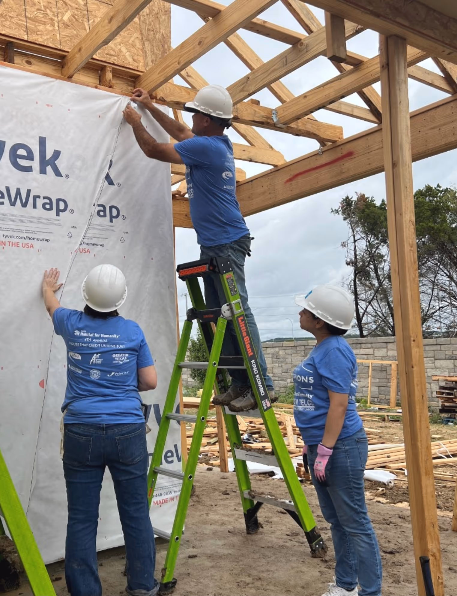 Three construction workers in blue shirts and white helmets installing Tyvek HomeWrap on a wooden house frame, with one worker on a green ladder.
