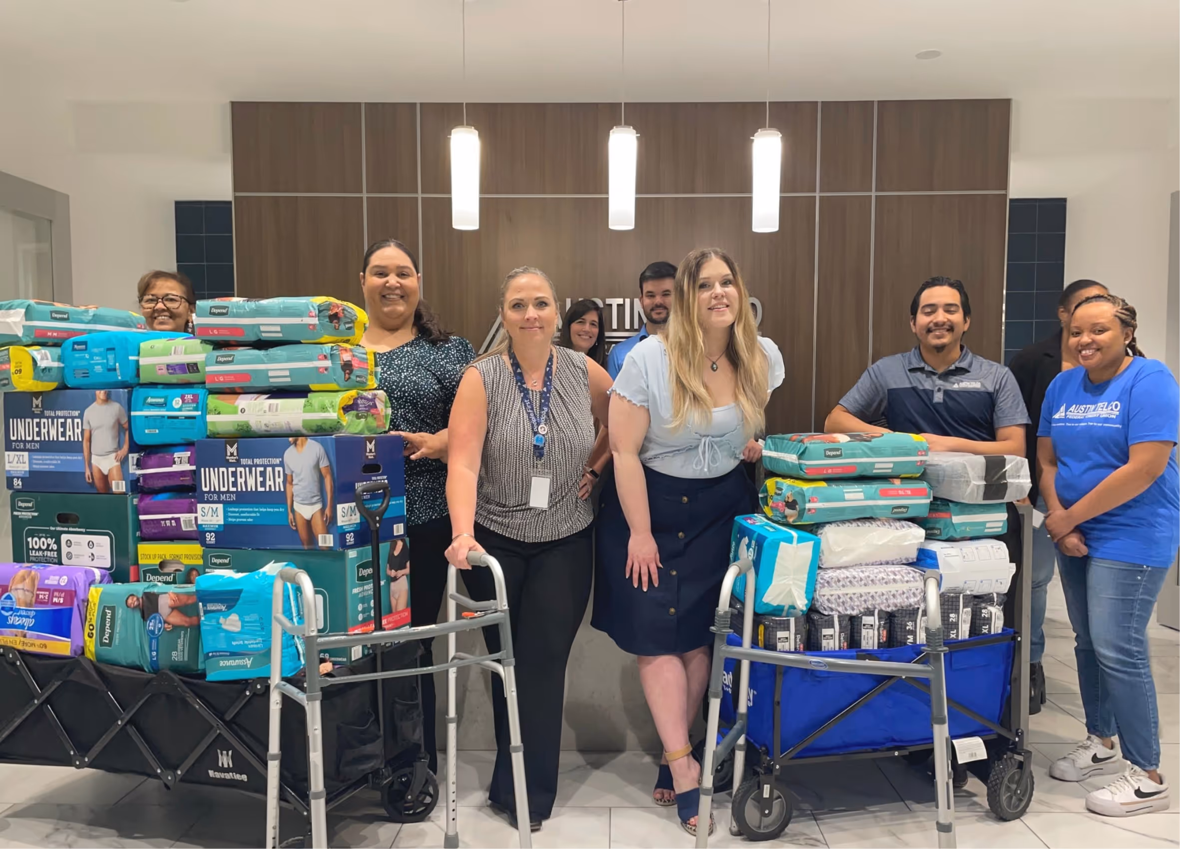 A diverse group of eight people standing behind two carts filled with packages of adult underwear and incontinence products in an indoor setting.