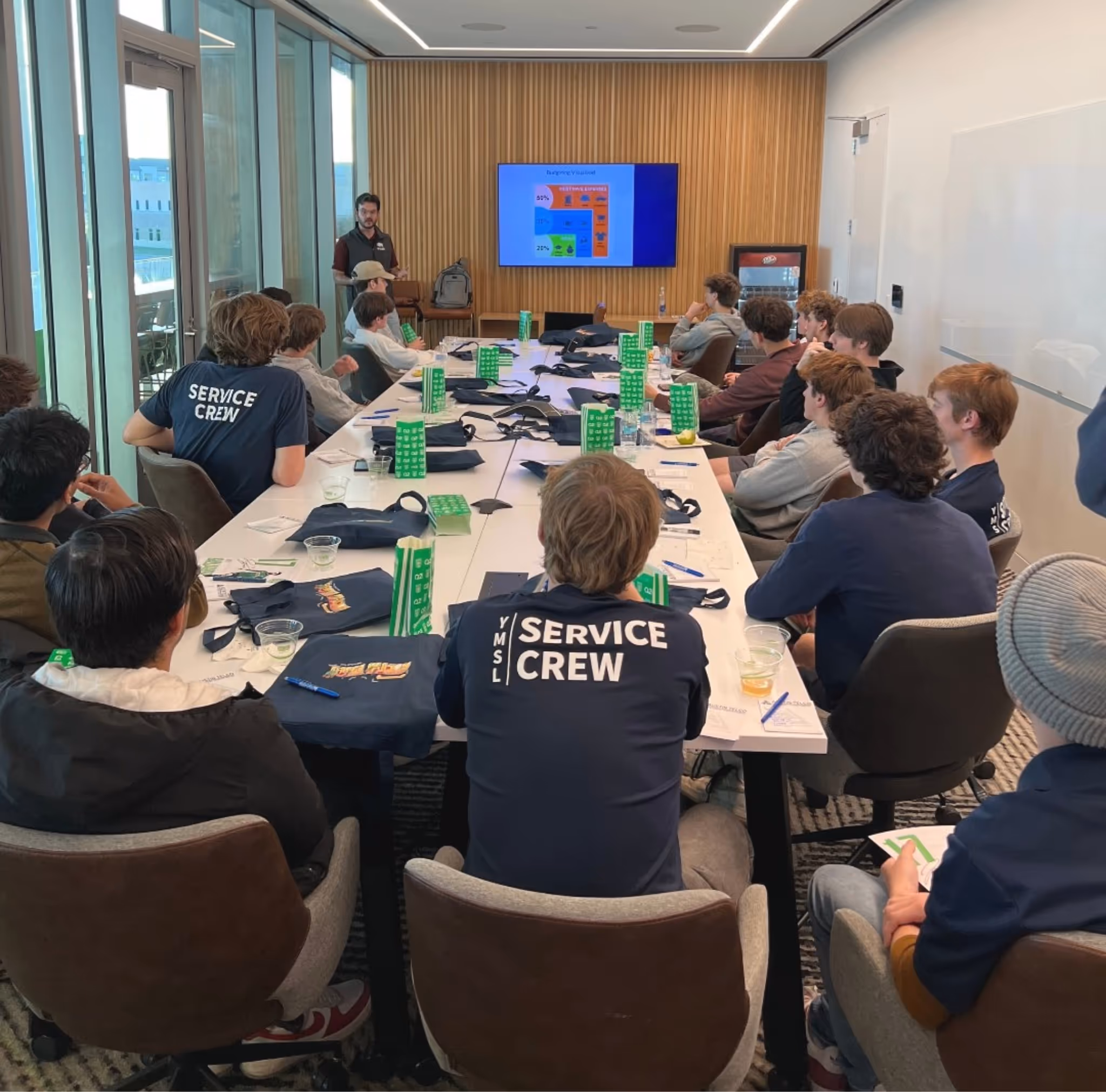 Group of young people seated around a conference table listening to a presenter standing near a screen with a colorful slide.