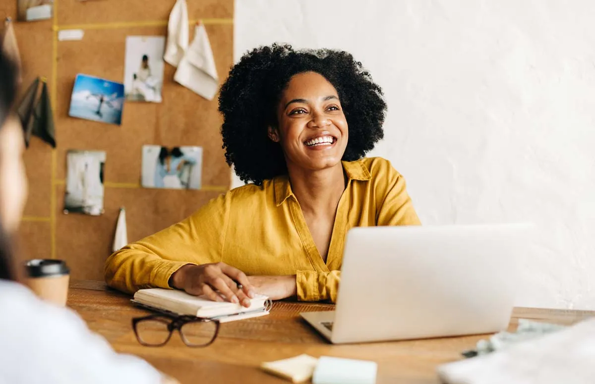 Smiling woman with curly hair in yellow shirt sitting at a table with laptop and notebook.