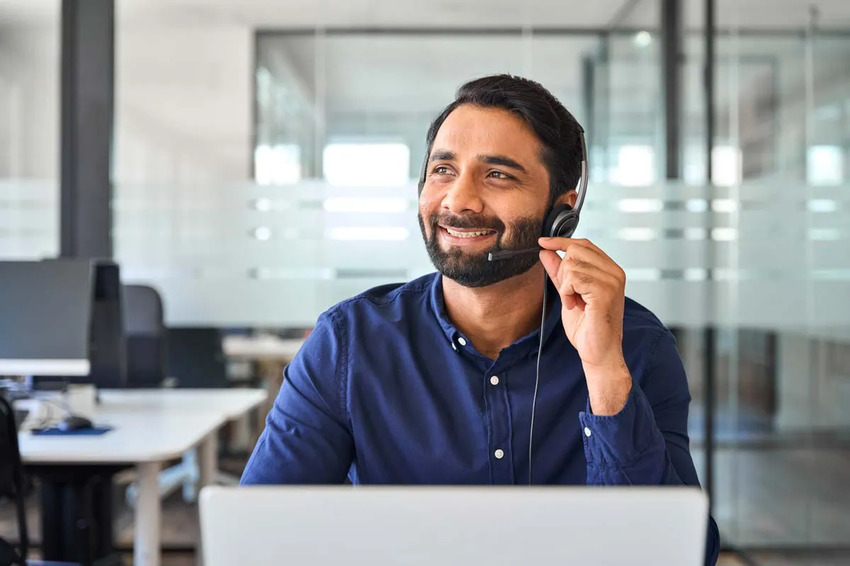 Smiling man wearing headset with microphone working at laptop in modern office.