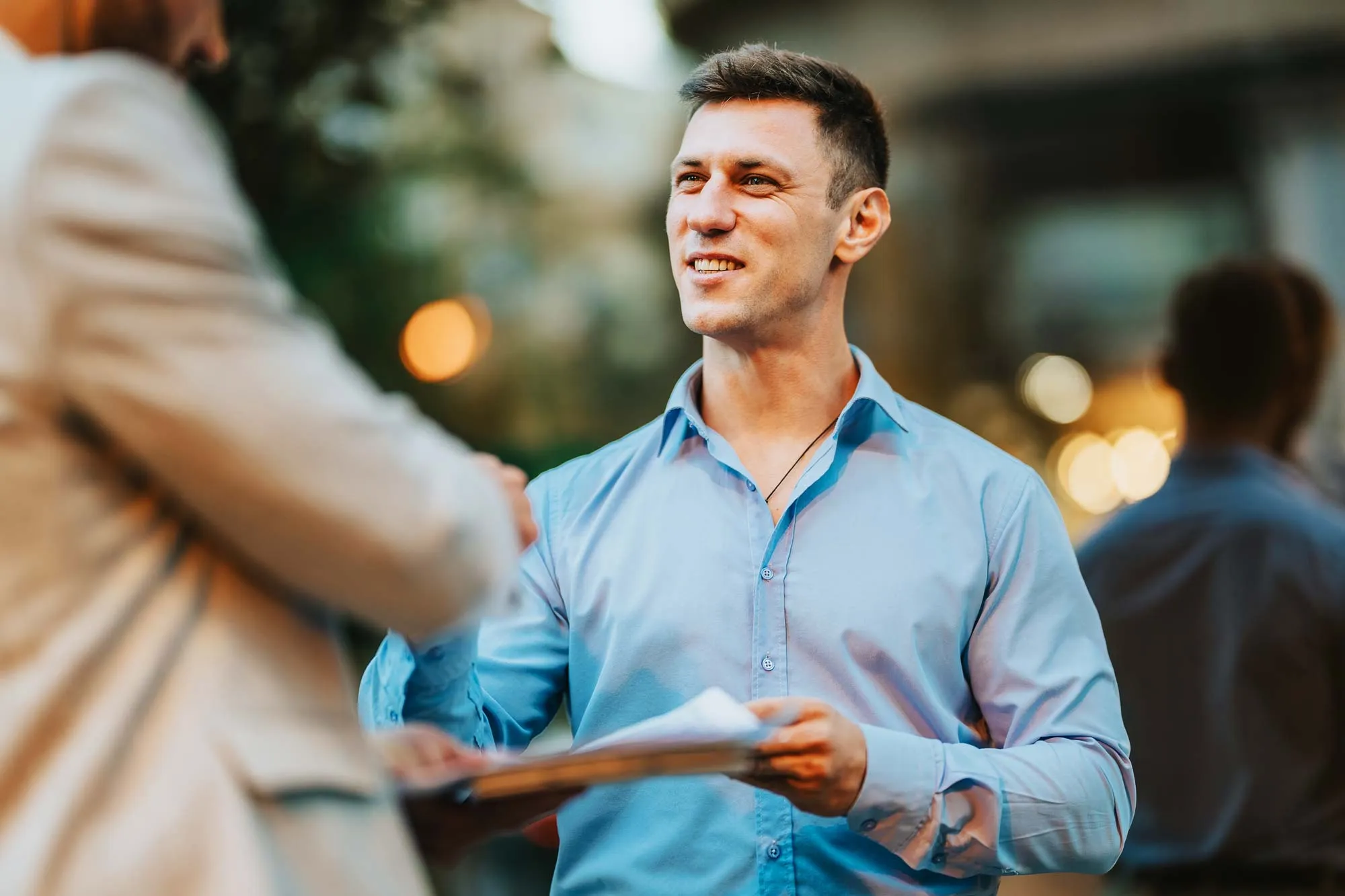 Man in blue shirt smiling and shaking hands while holding documents in an outdoor setting.