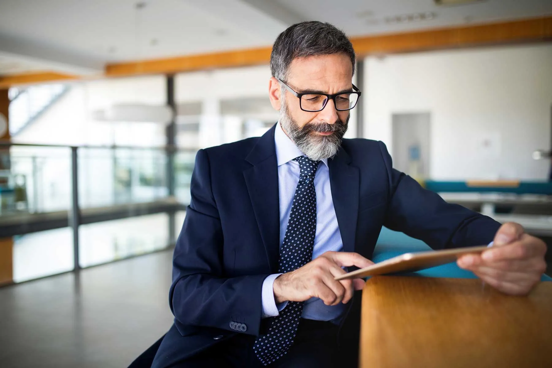 Mature man in suit and glasses using a tablet at a modern office desk.