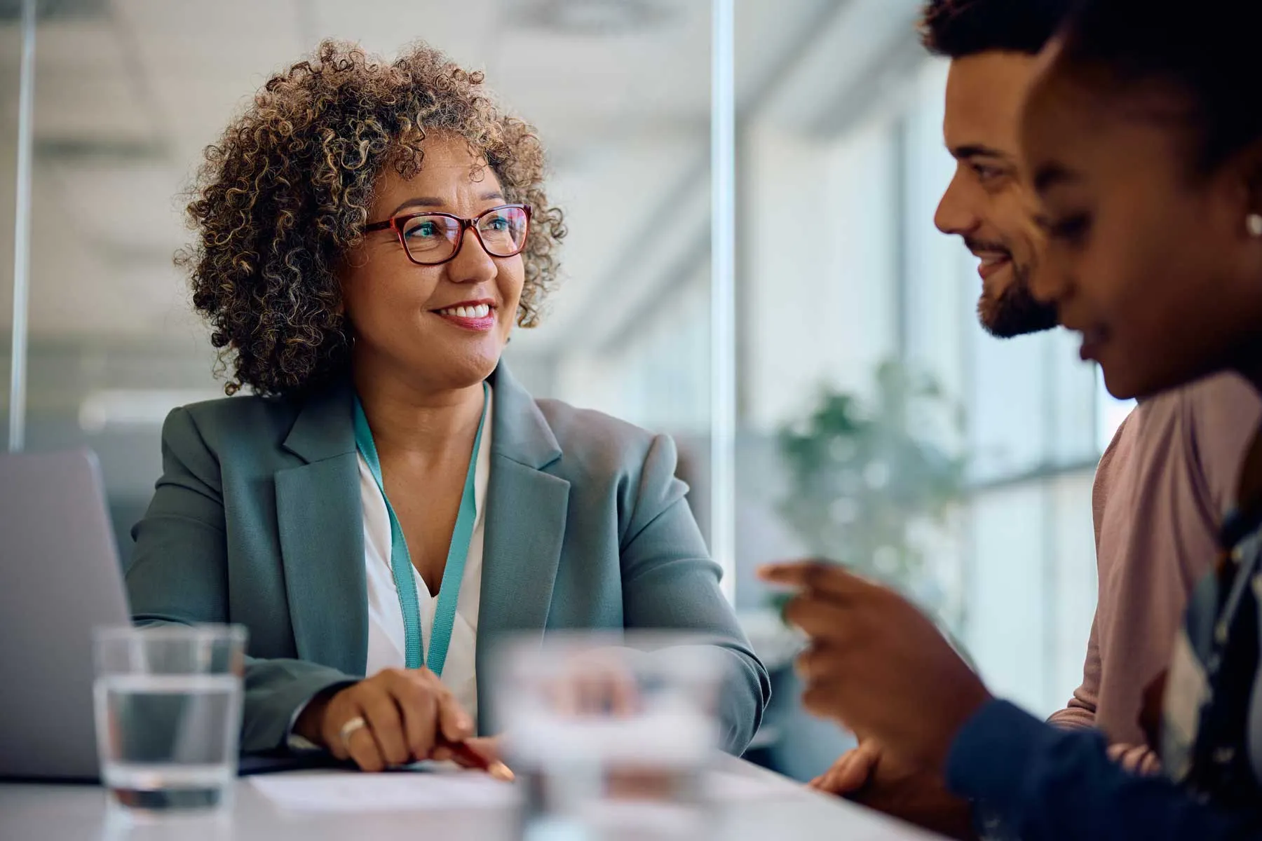 Smiling woman in glasses and blazer engaged in discussion with two colleagues at a table.