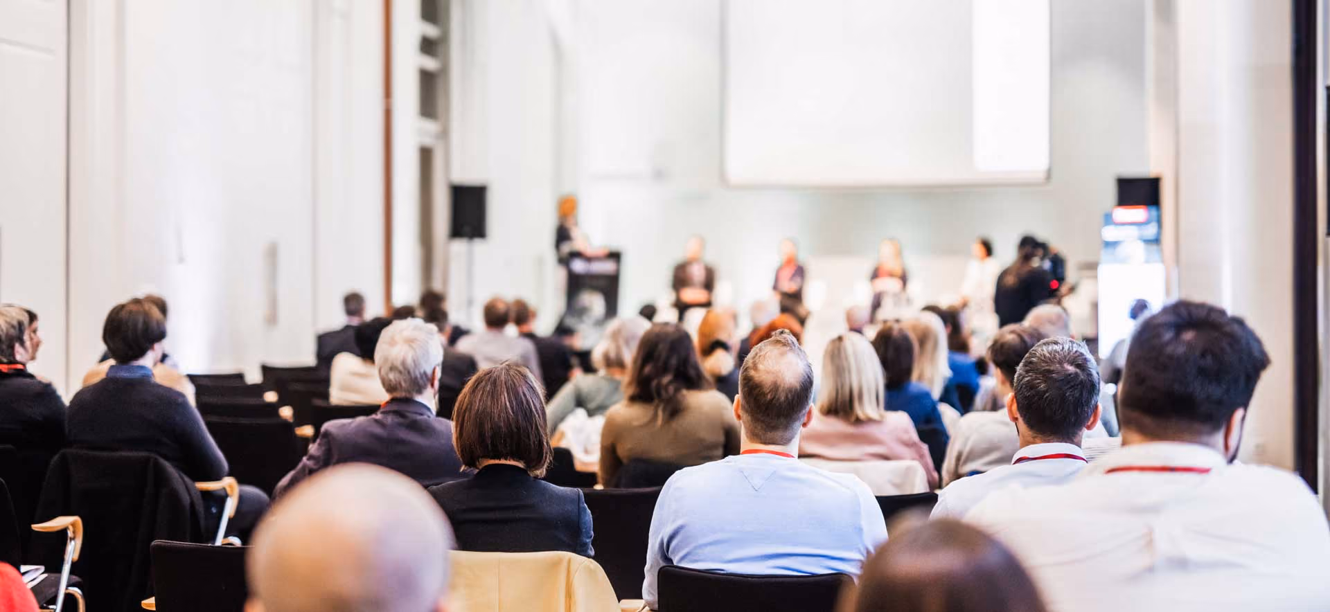 Audience seated facing a panel of speakers in a white conference room with a large screen.