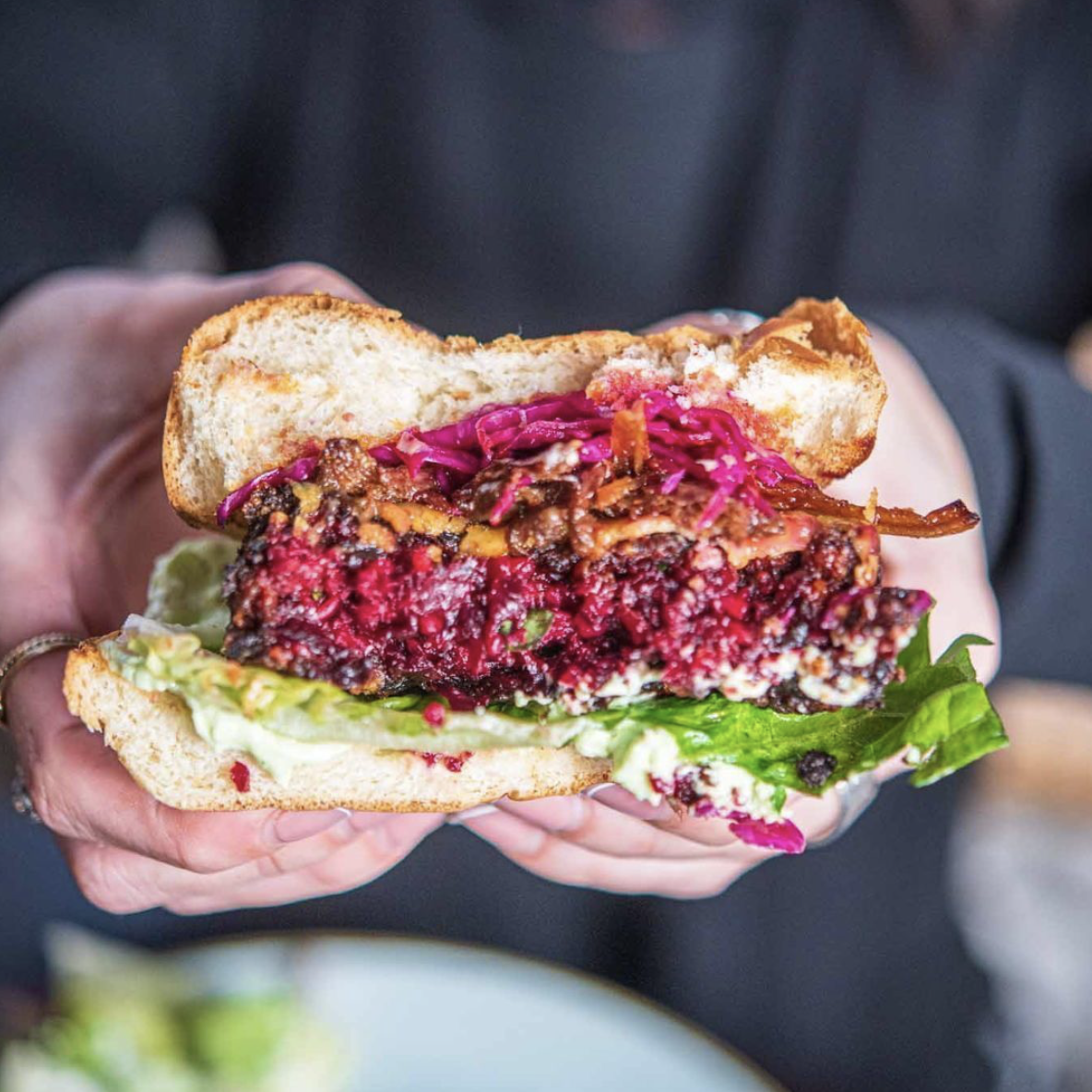 A photo of someone holding in their hands the vegan Beetroot Cheeseburger crispy lettuce, aioli, smoked applewood cheddar, bbq oyster mushrooms, pickled red cabbage.