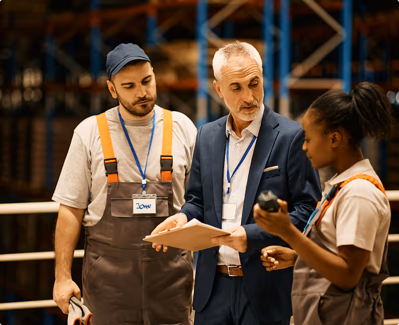 Three people in a warehouse setting, two in work overalls and one in business attire holding a clipboard