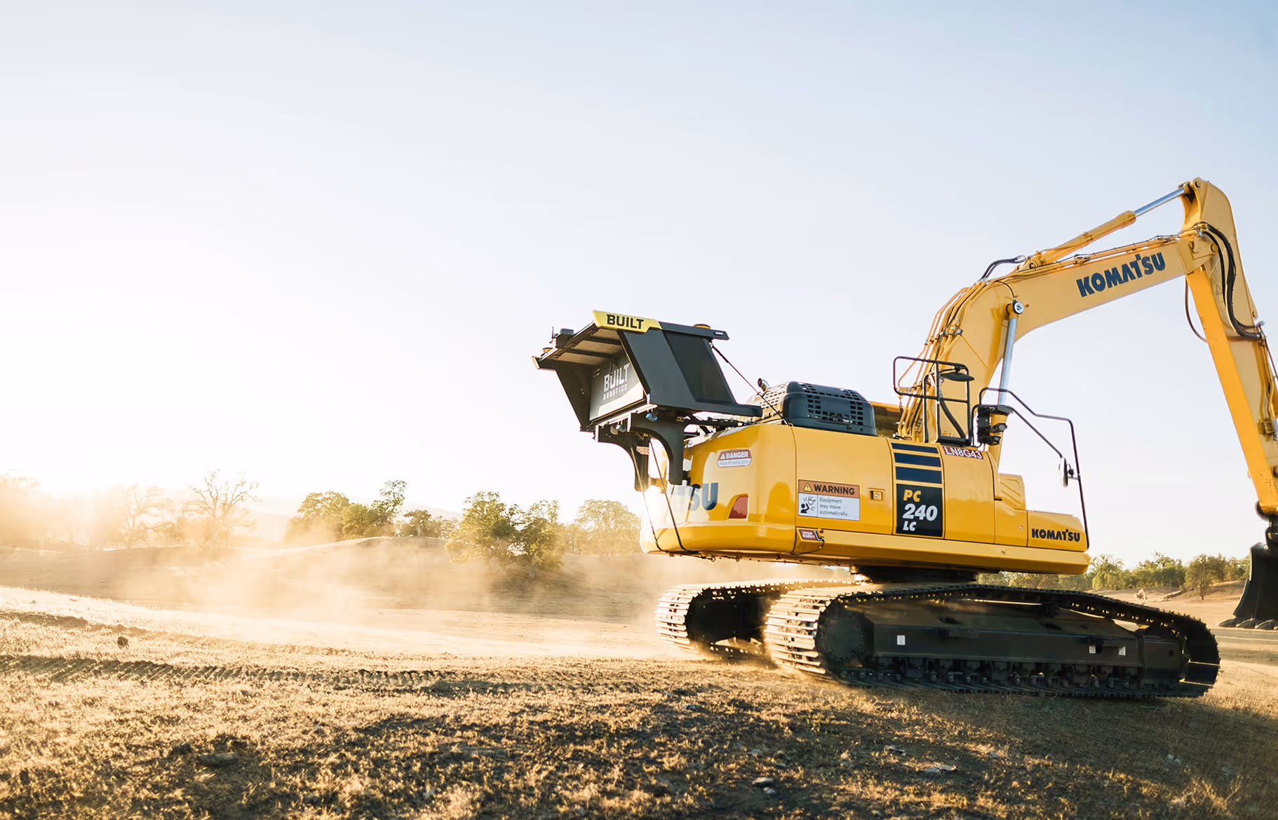 Excavator operating in a dusty field with trees and clear sky in the background.