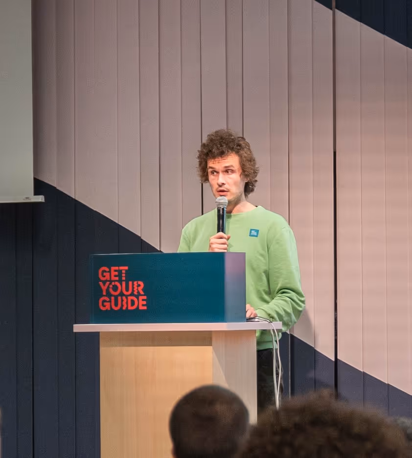 A man in a light green shirt is speaking at a podium in front of an audience, with a large screen behind him displaying a presentation titled "Intro" and bullet points for "Software Designs," "ML Designs," and "Conclusion."