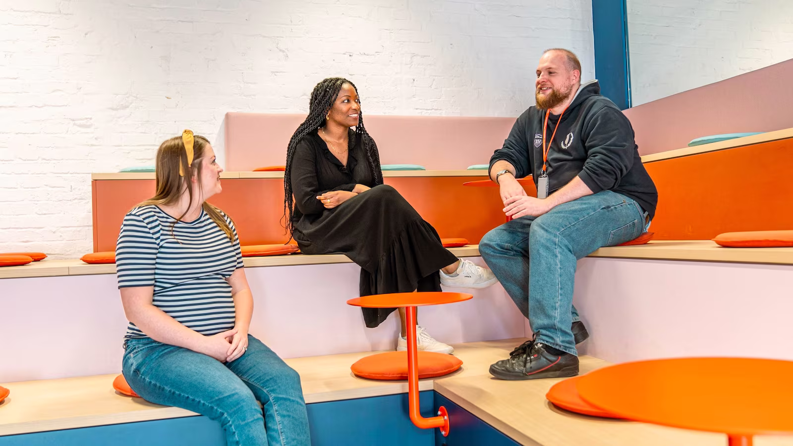 Three people are seated on tiered seating in a modern, brightly lit space. A man in a dark hoodie and jeans sits on the left, an African American woman in a black dress sits in the middle looking towards him, and a woman in a striped shirt and jeans sits on the right. An orange table is in the foreground.