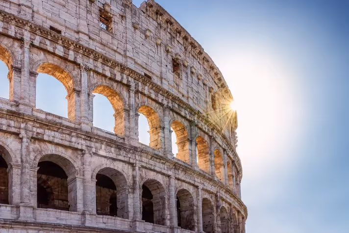 The ancient stone arches of the Colosseum in Rome are visible from a low angle, with bright sunlight shining through some of the upper arches against a clear blue sky.