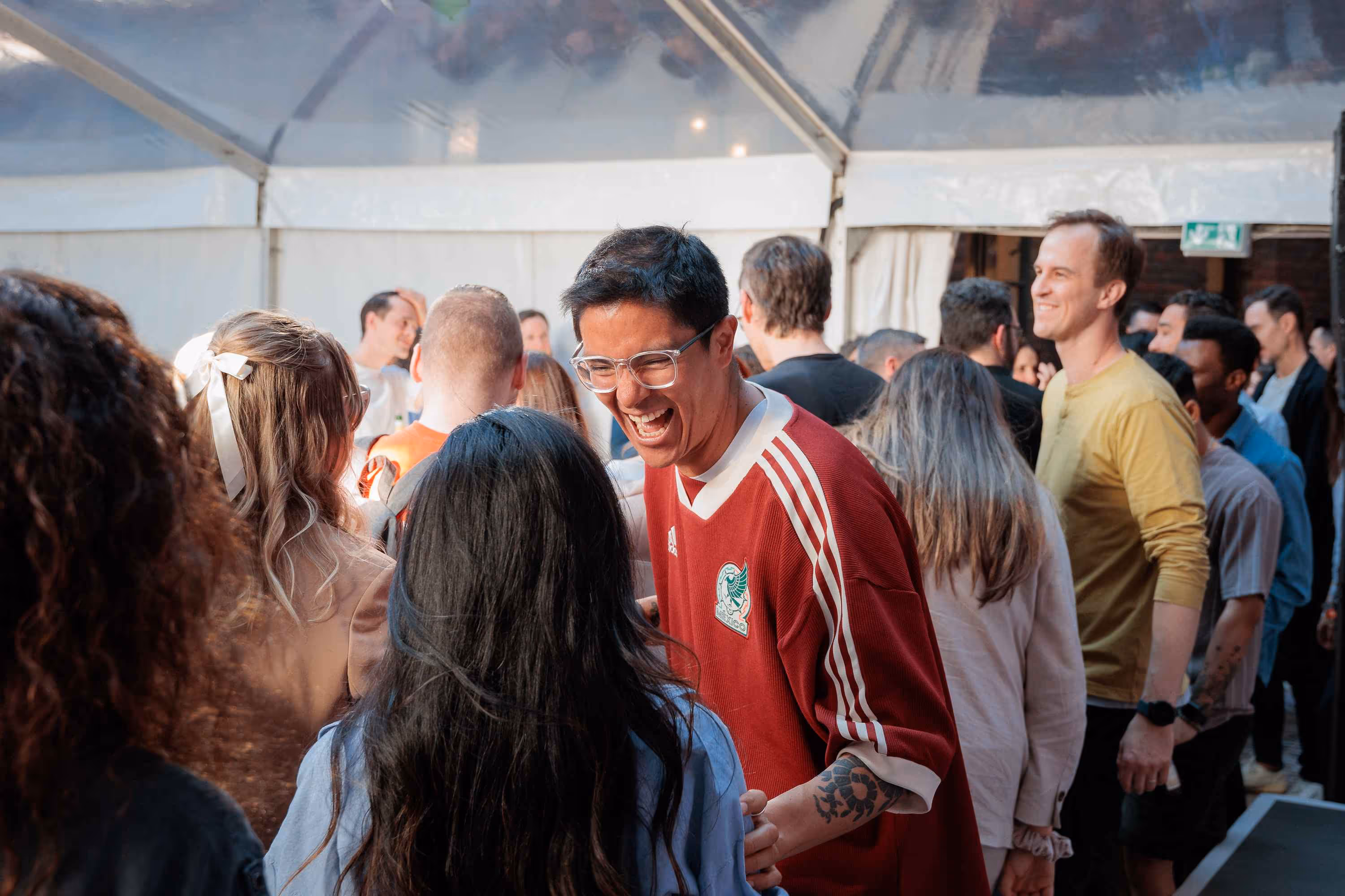 A man in a red and white striped shirt and glasses laughs heartily while looking at a person with long dark hair, who is seen from behind, in a crowded indoor setting.