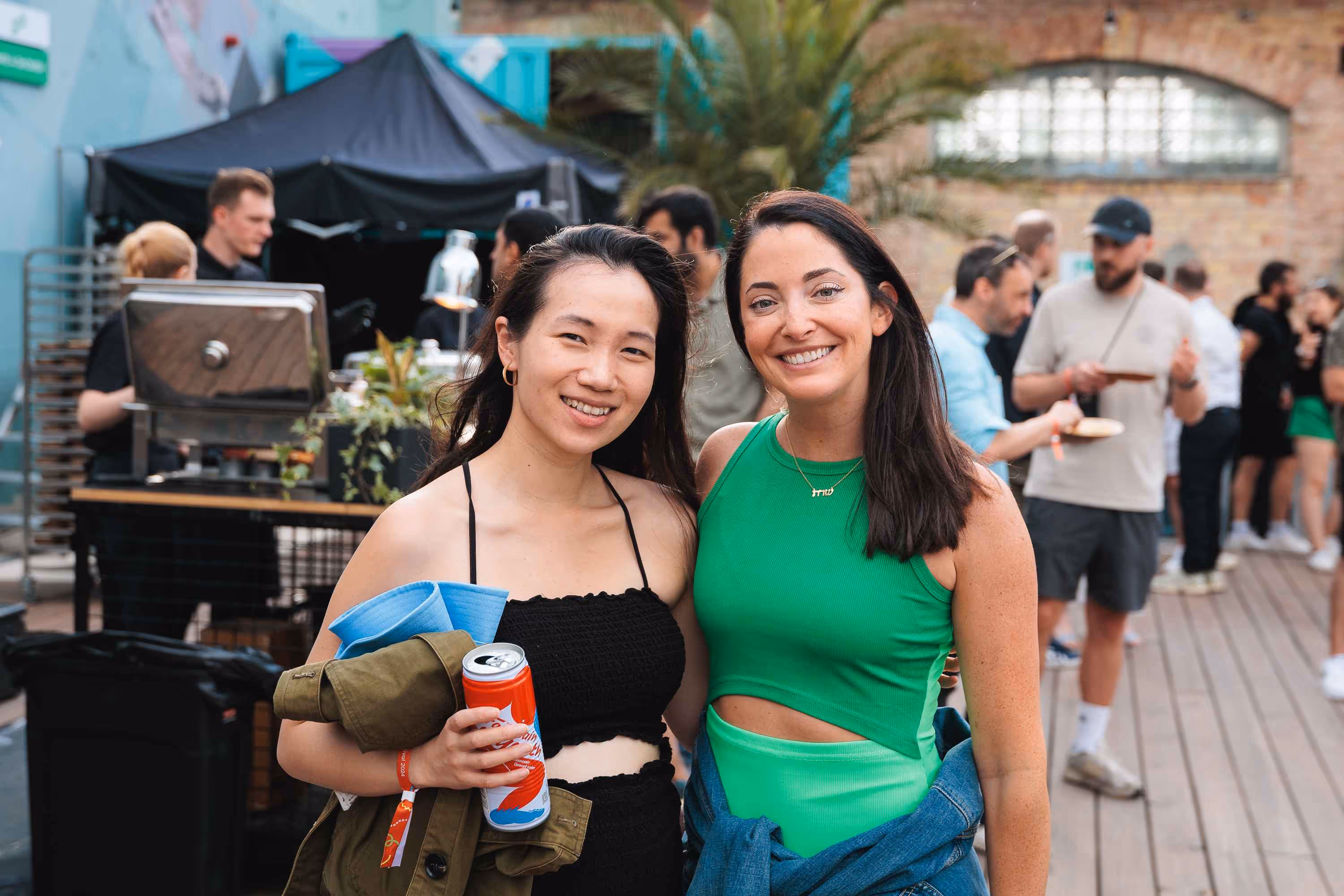 Two smiling women, one with dark hair holding a can and a jacket, and the other with long dark hair and a green top, pose for a photo at an outdoor event with blurred people and a BBQ station in the background.