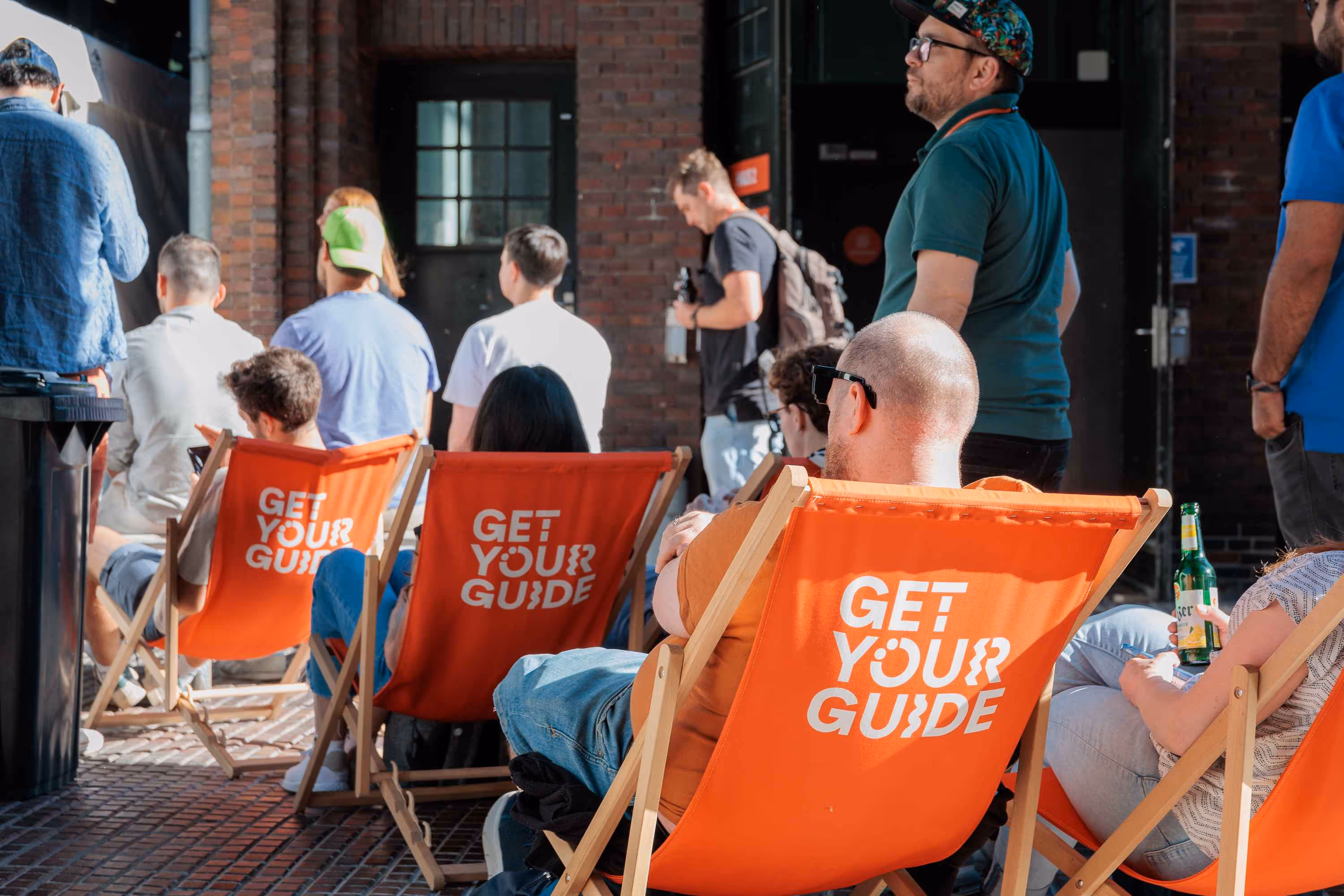 Several people are seated outdoors in orange canvas sling chairs that say "GET YOUR GUIDE" on the back. They are facing away from the camera, looking towards a building and other blurred people in the background.