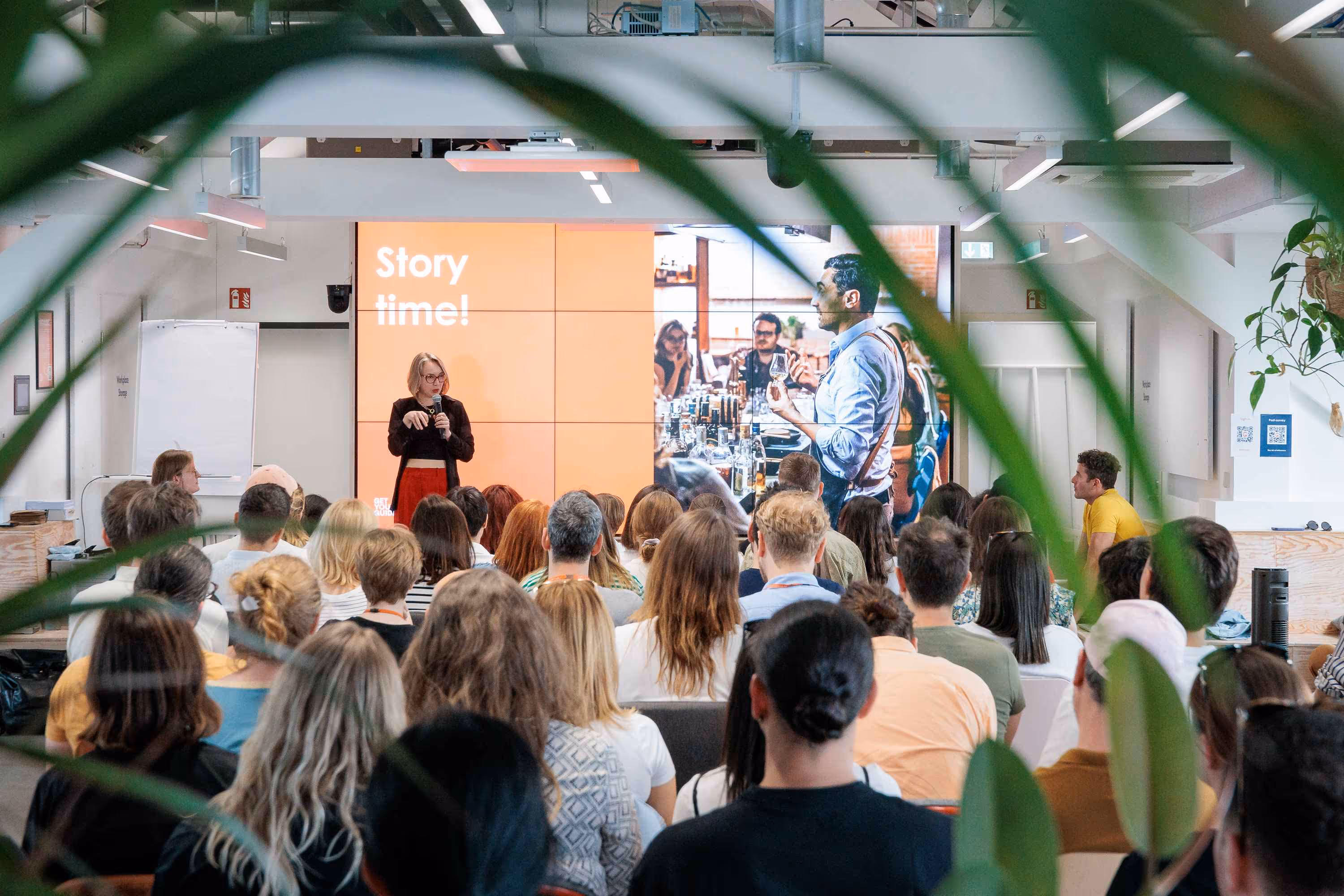 A speaker stands on a stage in front of a large screen displaying "Story time!" and an image of people, addressing a seated audience from behind.