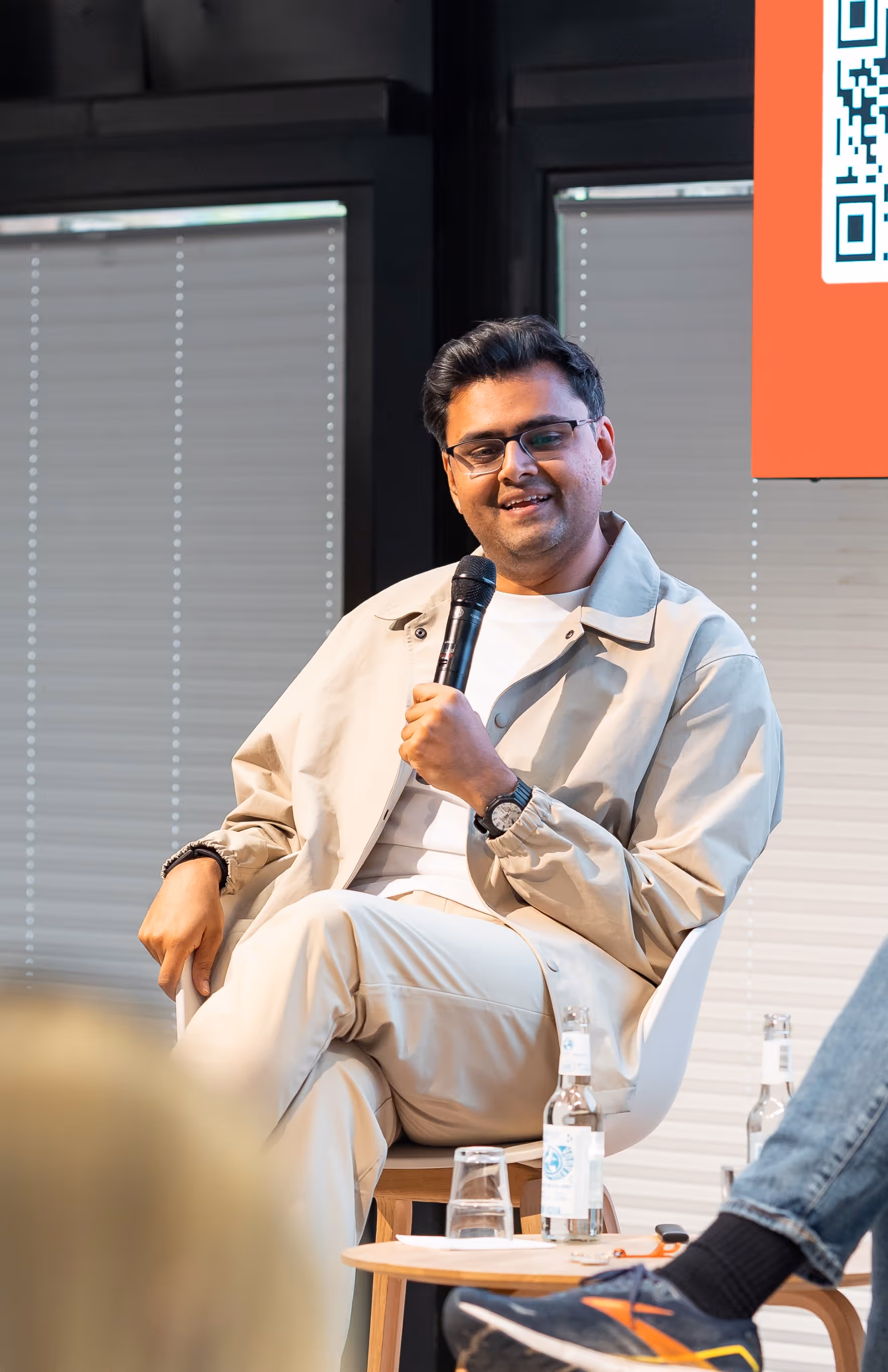A man with glasses and a light-colored jacket smiles and gestures with one hand while seated on a stool, holding a microphone. A blurred QR code is visible in the background.