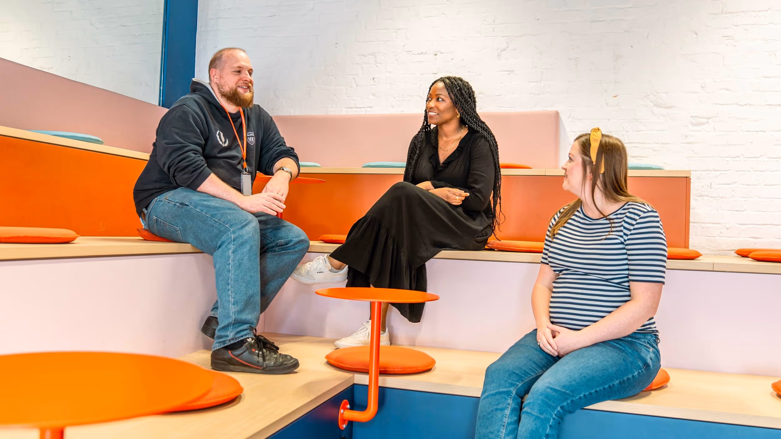 Three people are seated on tiered seating in a modern, brightly lit space. A man in a dark hoodie and jeans sits on the left, an African American woman in a black dress sits in the middle looking towards him, and a woman in a striped shirt and jeans sits on the right. An orange table is in the foreground.