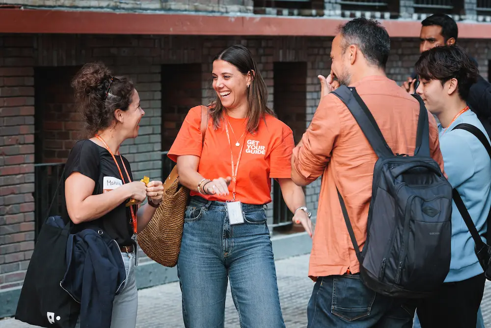 A woman wearing a orange get your guide shirt while talking to the other women in black. Other men wearing bags are also listening to the conversation