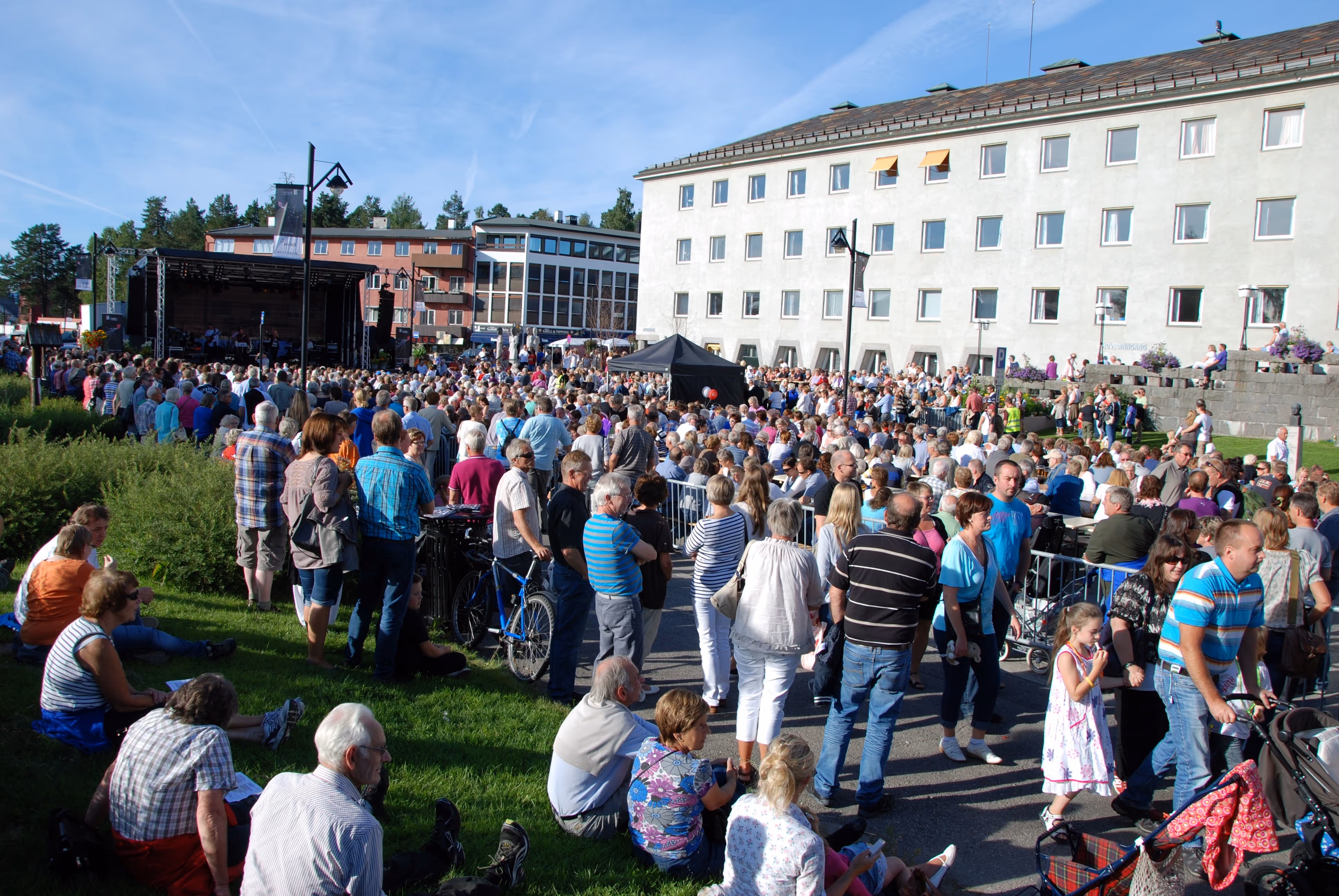 Stor folkemengde samlet utendørs foran en scene under en solrik dag i et byområde med hvite og røde bygninger.