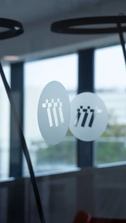 Due adesivi tondi opachi con un logo stilizzato bianco a forma di tre figure con quadrati sopra, attaccati su una superficie trasparente con sfondo di finestre.