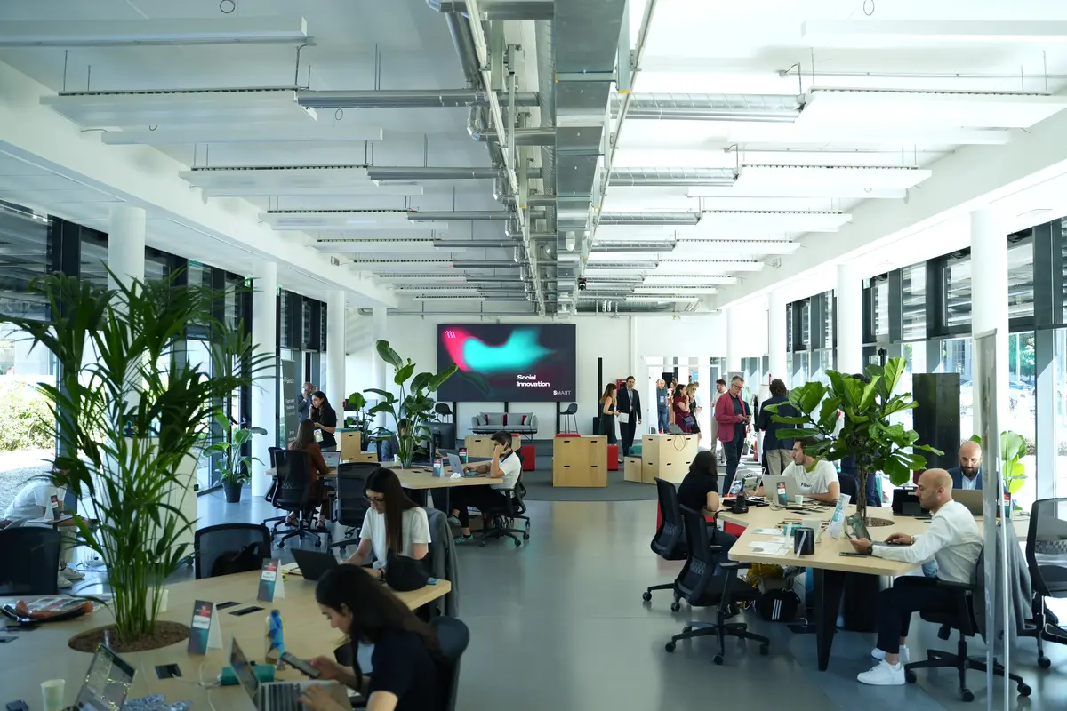 Ufficio moderno open space con persone che lavorano a computer e piante verdi tra le scrivanie.