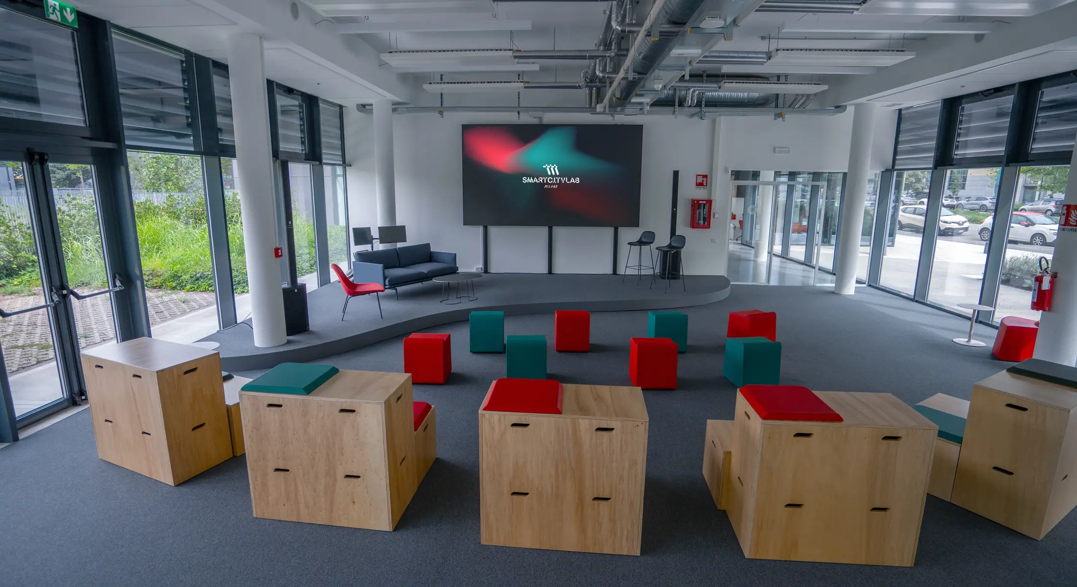 Spazio moderno con sedute colorate in legno e tessuto, grande schermo con logo Smart City Lab su parete bianca e ampie finestre con vista sul parcheggio e il verde esterno.