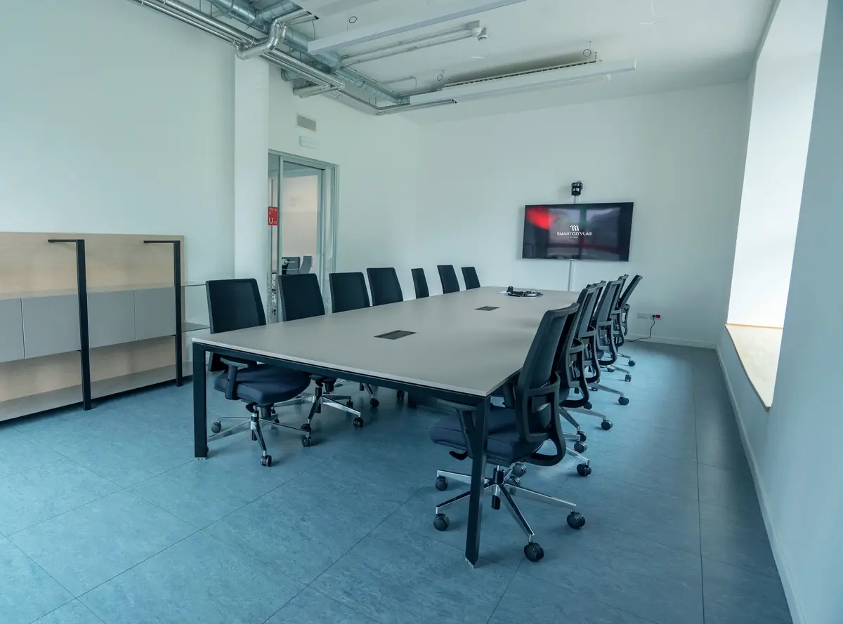 Sala riunioni moderna con tavolo rettangolare grigio, dodici sedie girevoli nere e schermo TV a muro con logo SmartCityLab.