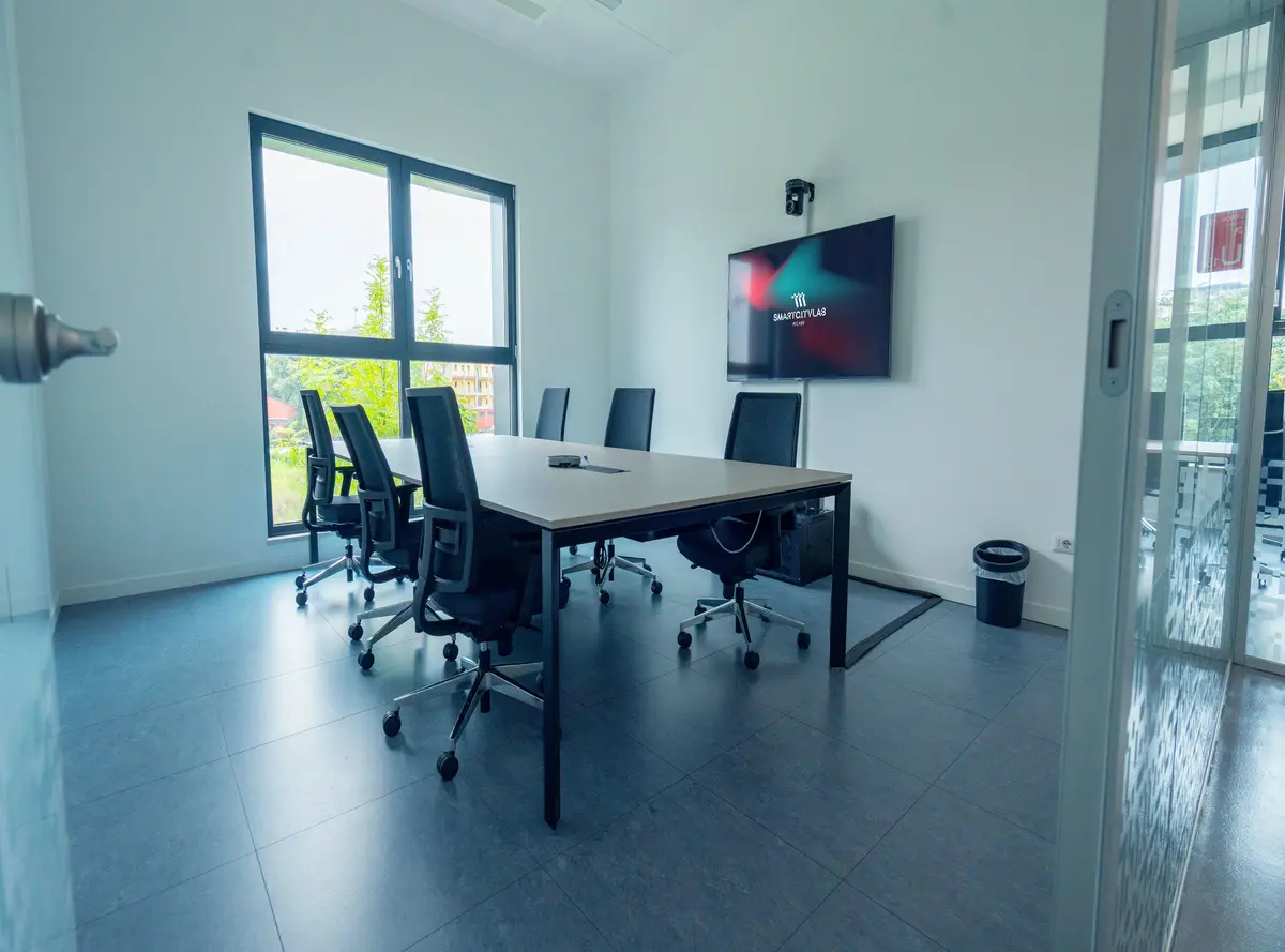 Sala riunioni moderna con sei sedie nere girevoli intorno a un tavolo rettangolare e una TV a schermo piatto montata sulla parete bianca.