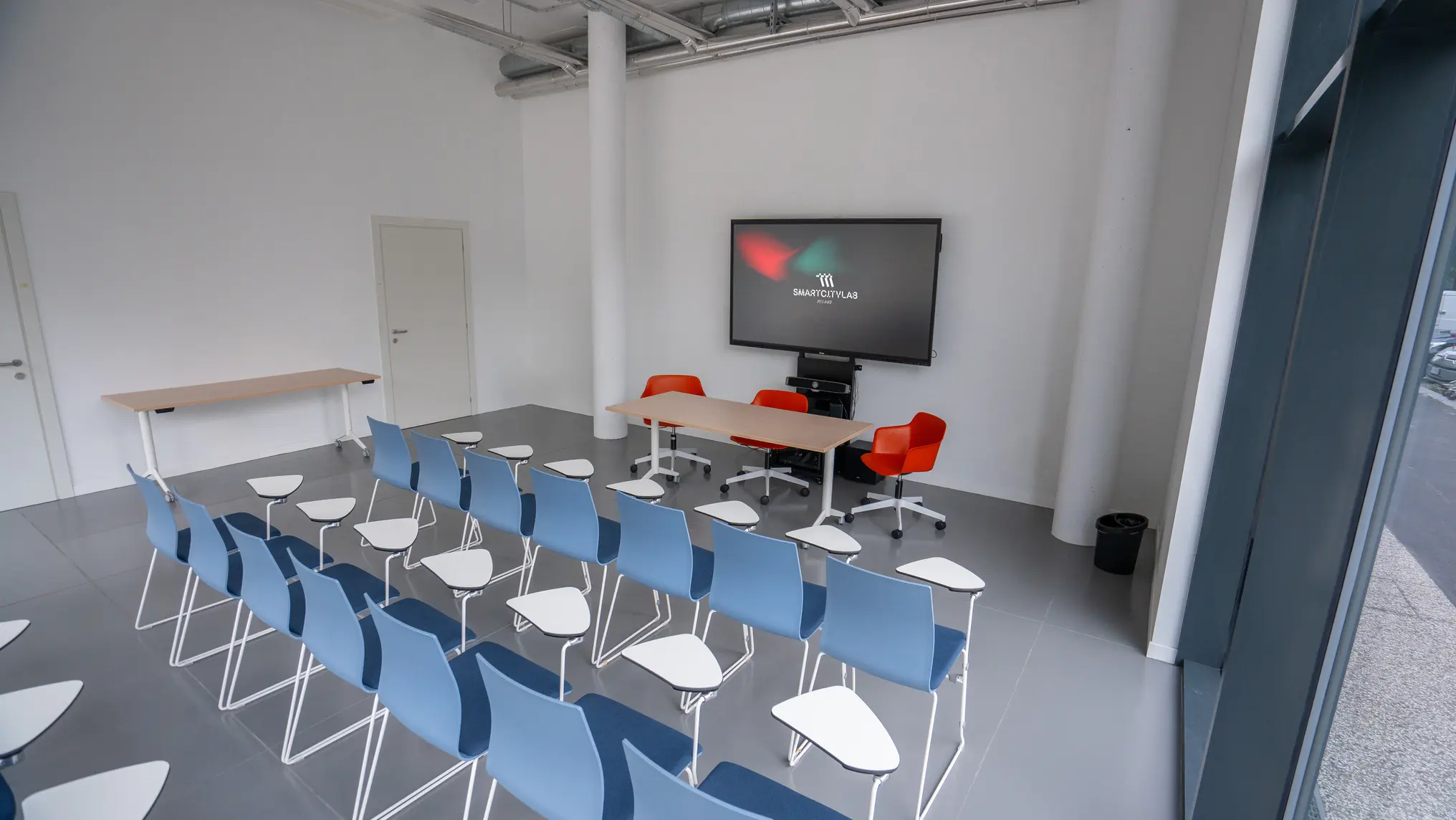 Aula moderna vuota con file di sedie blu dotate di tavoletta per scrivere, tre sedie rosse e un grande schermo con logo Smartcitylab.