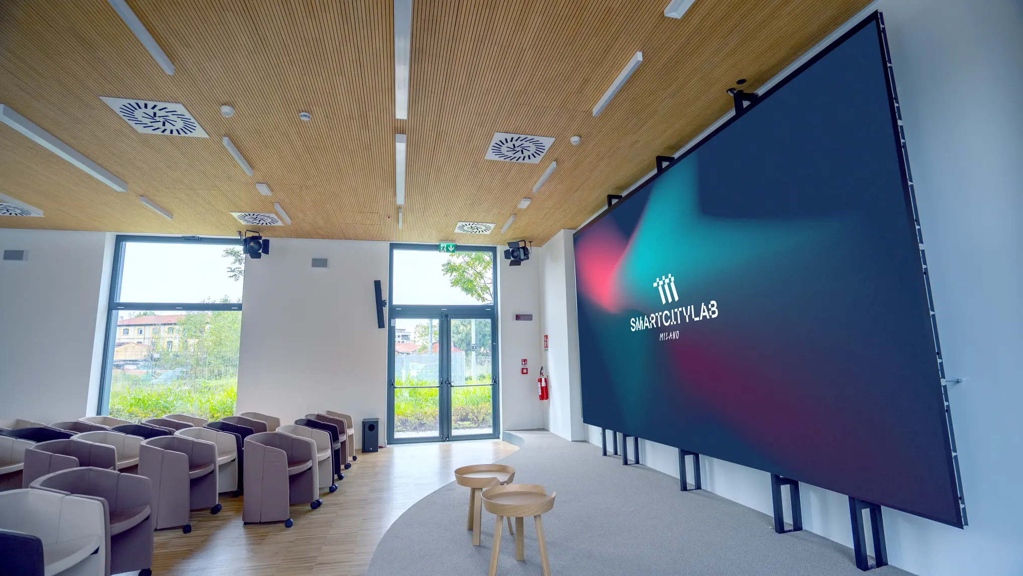 Sala conferenze moderna con sedie disposte in file, parete con grande schermo che mostra il logo Smart City Lab Milano e finestre con vista esterna.