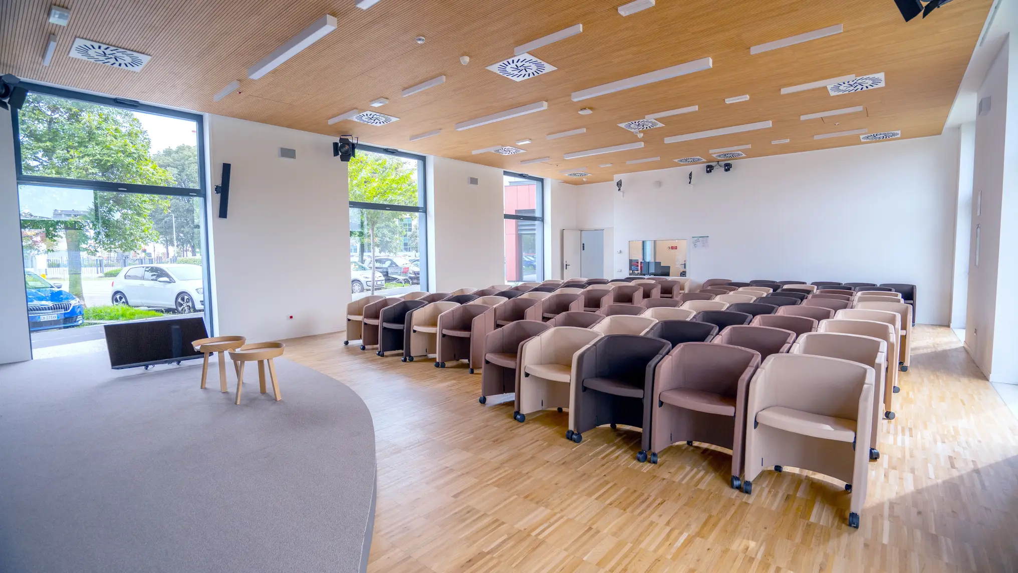 Sala conferenze moderna con file ordinate di poltrone mobili beige, marroni e nere e un palco con due tavolini di legno.
