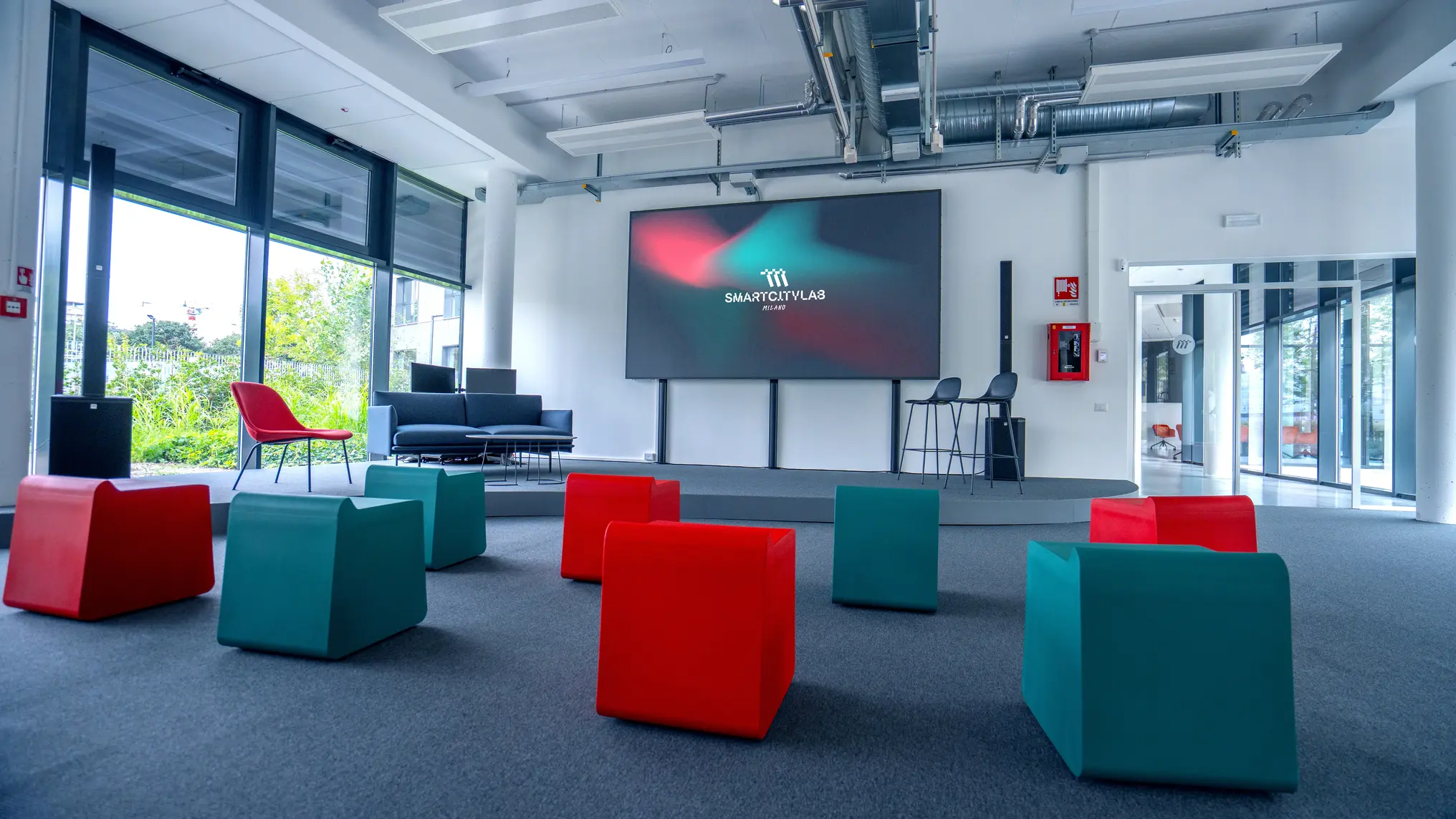 Sala conferenze moderna con schermo grande, sedie rosse e blu, e poltrone nere vicino a una vetrata.