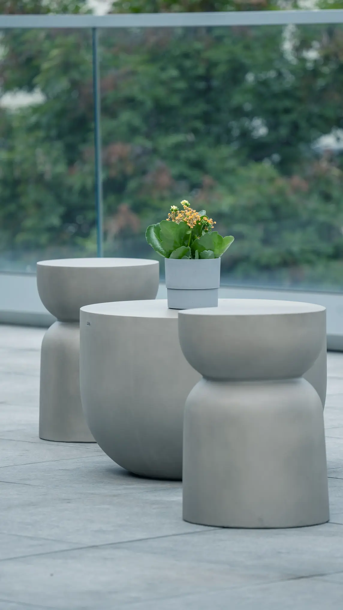 Tavolo e sgabelli moderni in cemento grigio con una pianta in vaso bianco sul tavolo, su un terrazzo con ringhiera in vetro e sfondo di alberi sfocati.
