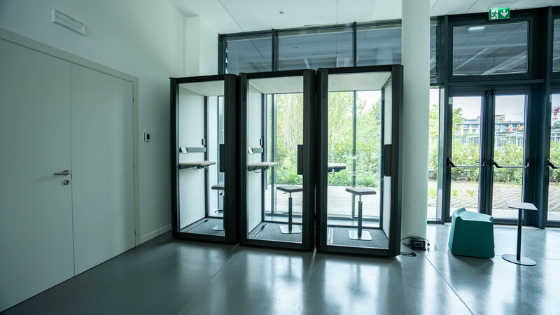 Tre cabine di telefonia moderne con sgabelli all'interno in uno spazio ufficio con grandi finestre e pavimento in cemento.