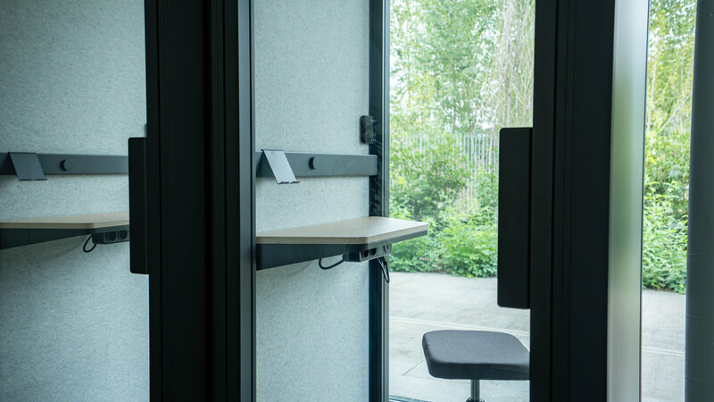 Postazione di lavoro minimalista con scrivania in legno, sedia grigia e pareti in tessuto grigio, affacciata su un'area verde esterna.