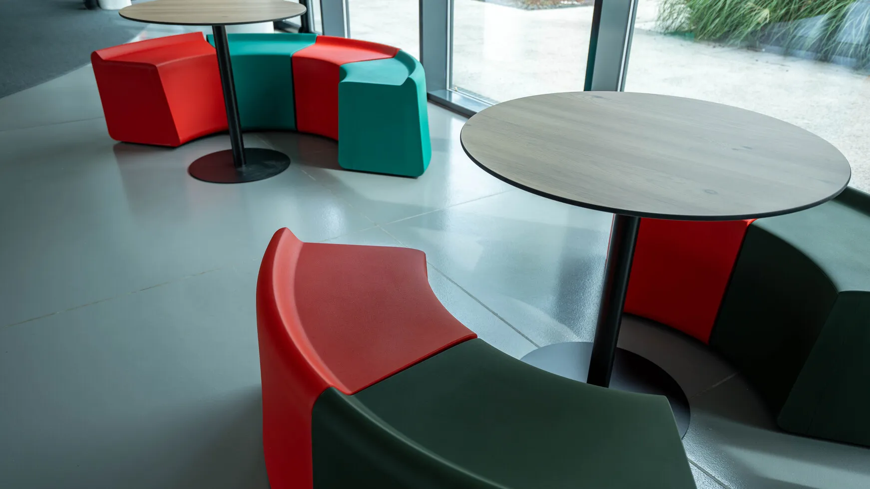 Sedute modulari rosse, verdi e turchesi attorno a tavoli rotondi in una stanza con pavimento grigio e grandi finestre.